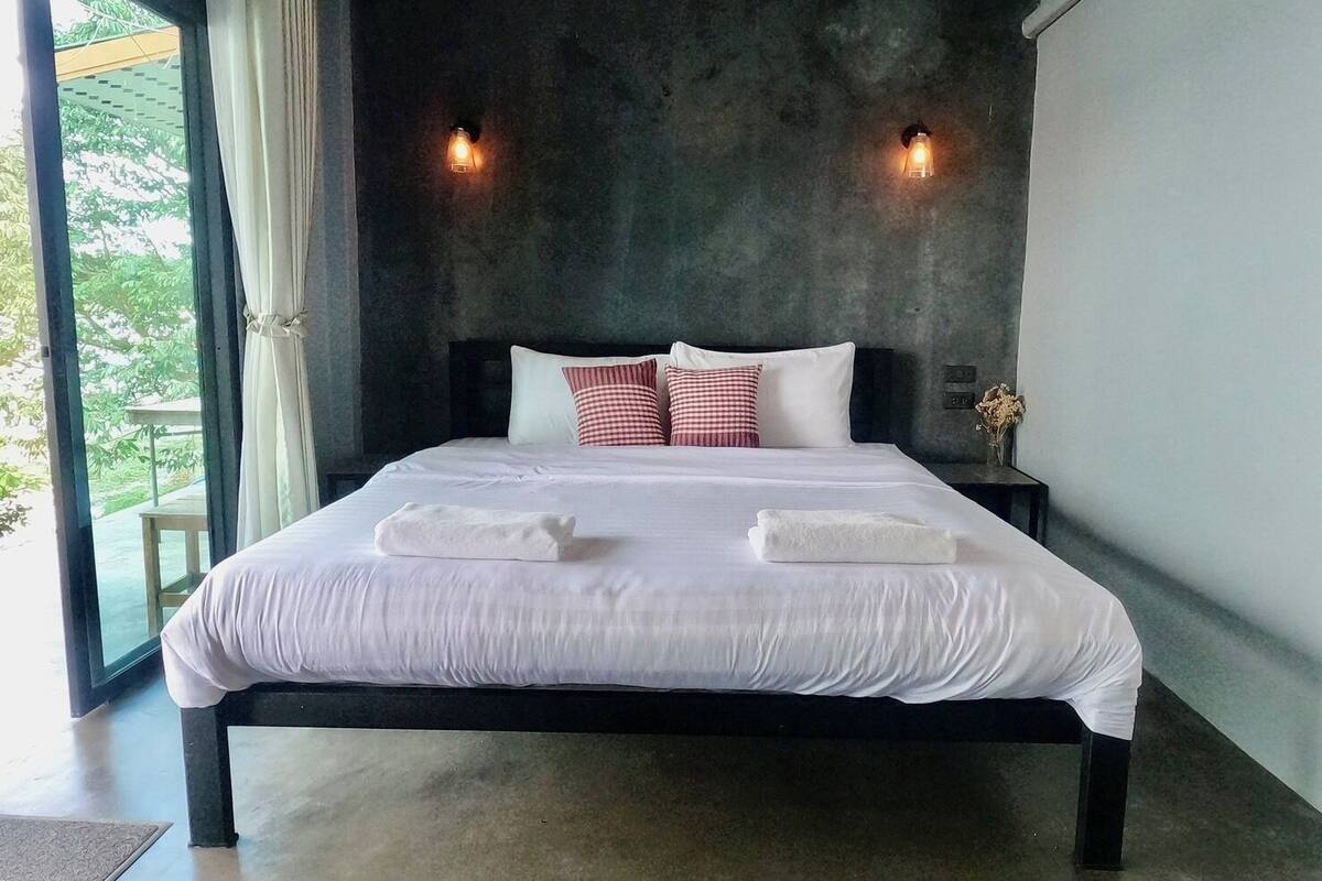 A modern bedroom with a large bed, two bedside lamps, and a view of greenery outside.