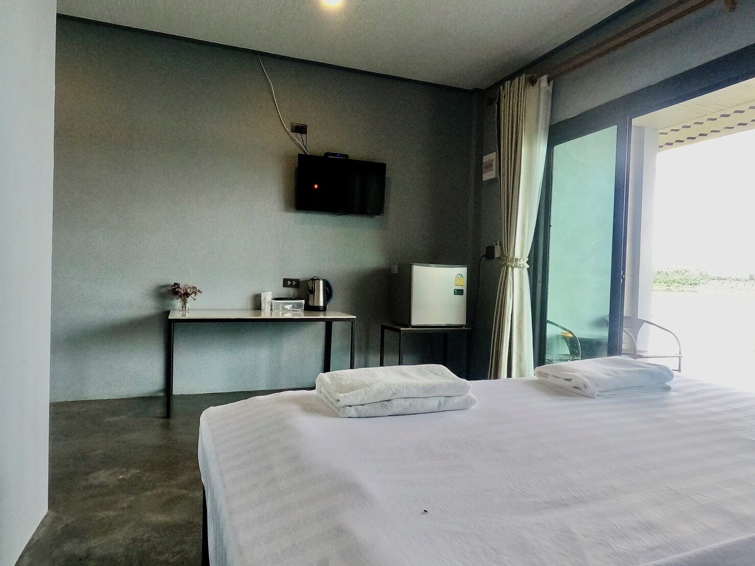 A hotel room with a bed, a small table with a kettle, a TV, and a refrigerator.