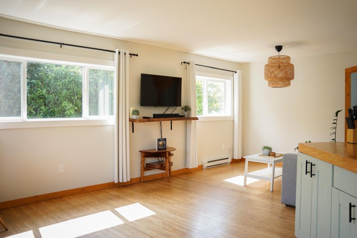 Ett rum med en platt-TV som är monterad på väggen, ett litet träbord, ett vitt skåp och en hängande lampa.