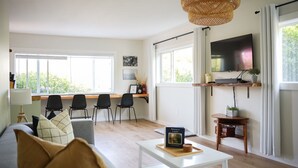 Un salon moderne avec un canapé, une table basse et un téléviseur fixé au mur. On y trouve trois chaises noires et un bureau avec un ordinateur.