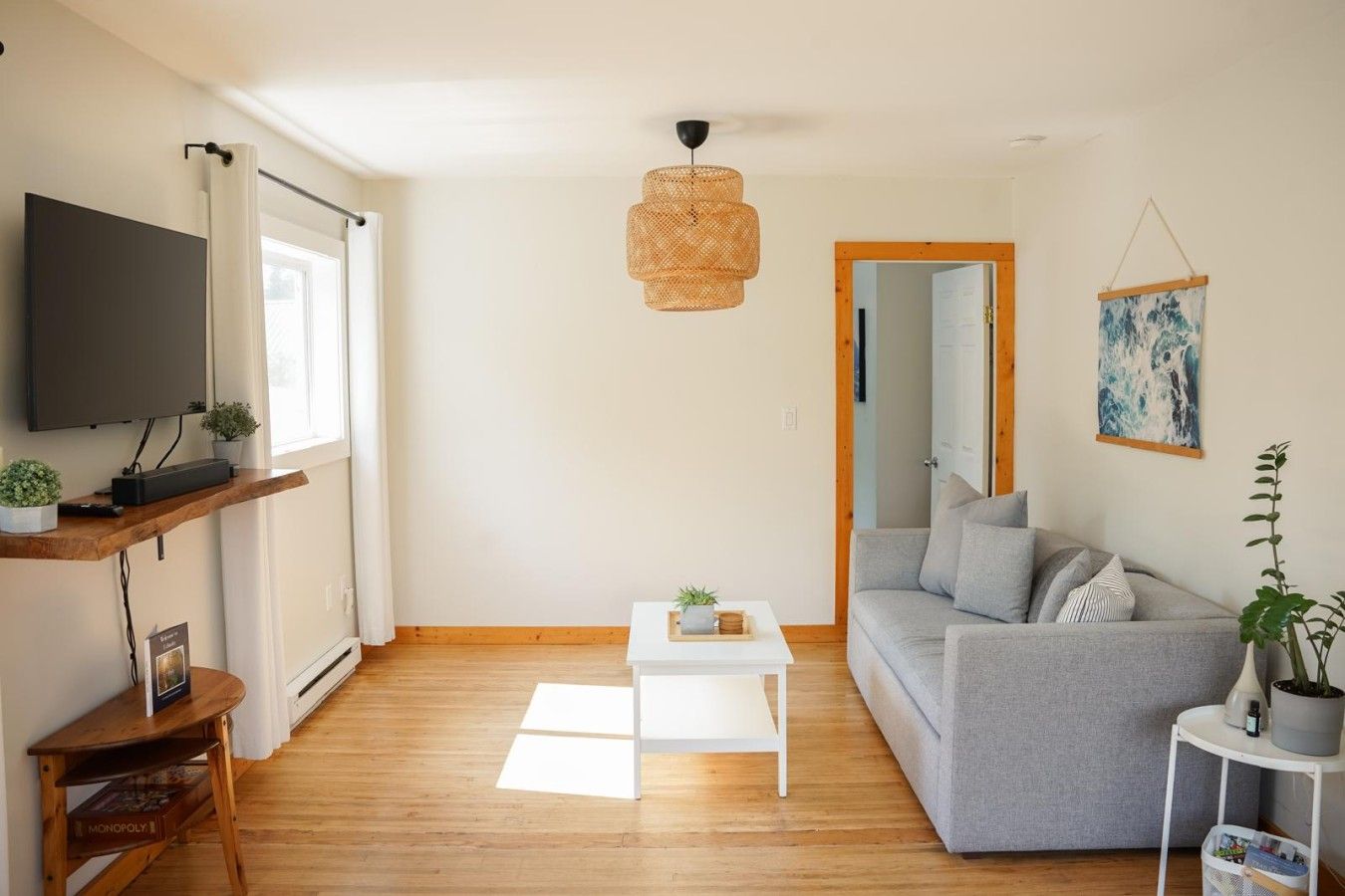 Una sala de estar con un sofá gris, una mesa de centro blanca, un televisor en un estante de madera, una lámpara colgante de fibras naturales y un cuadro en la pared.