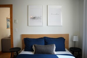 A hotel room with a bed, two framed pictures on the wall, a bedside lamp, and a small table.