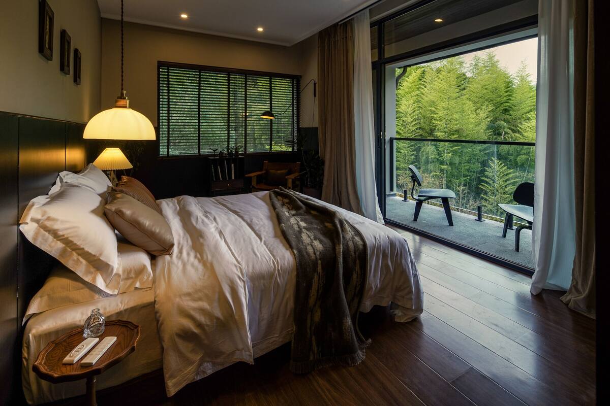A hotel room with a large bed, a bedside table with a lamp, a view of a balcony with outdoor seating, and a window with blinds.
