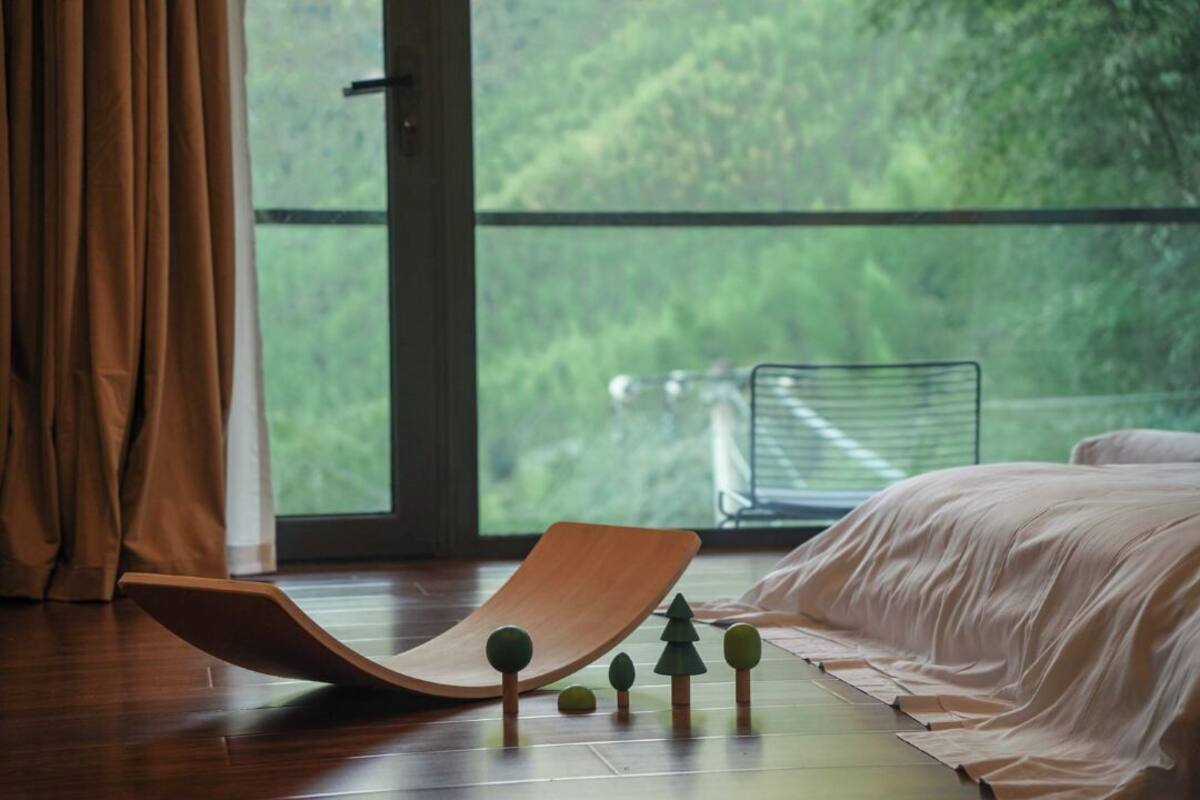A bedroom with a wooden chair, a bed, and a view of greenery through a window.
