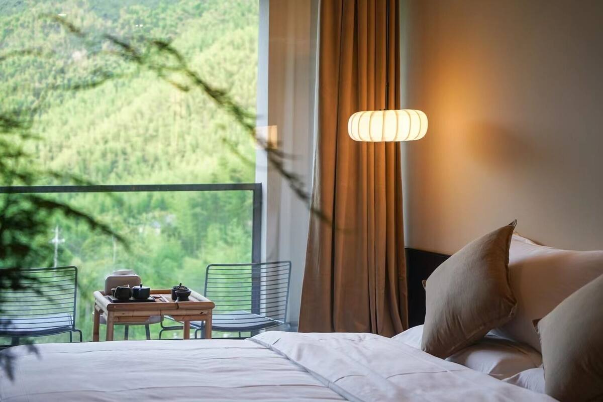A hotel room with a bed, a table with tea set, a chair, and a view of a forest through a window.
