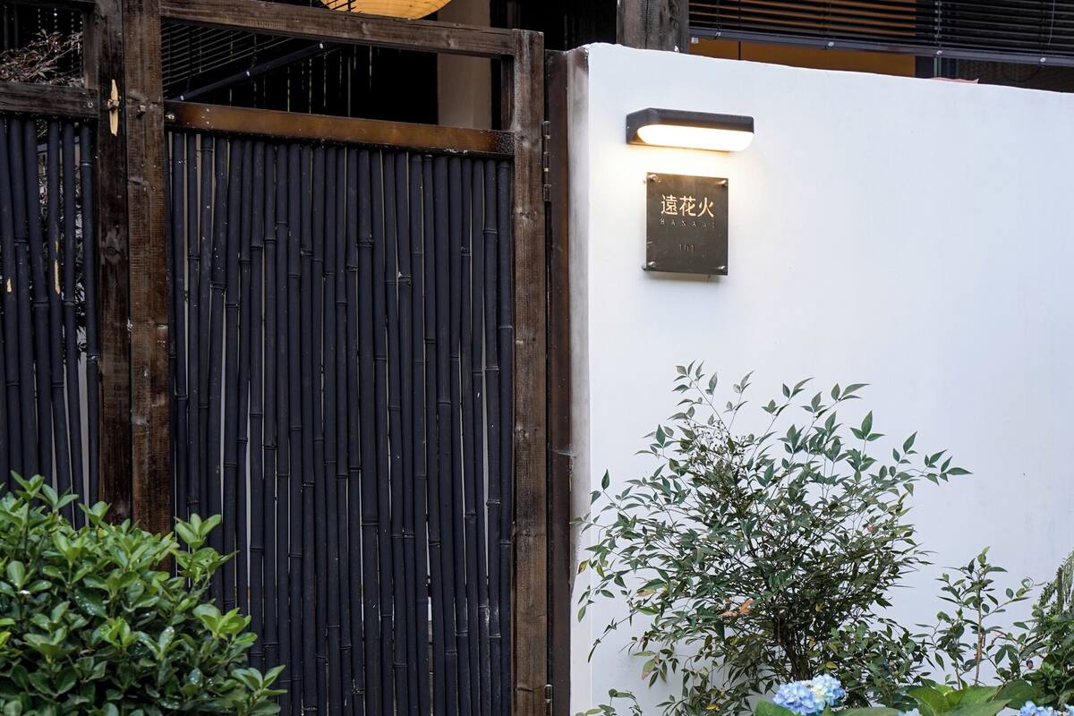 A traditional Japanese gate with a lantern above, a wooden fence, and a wall with a sign.