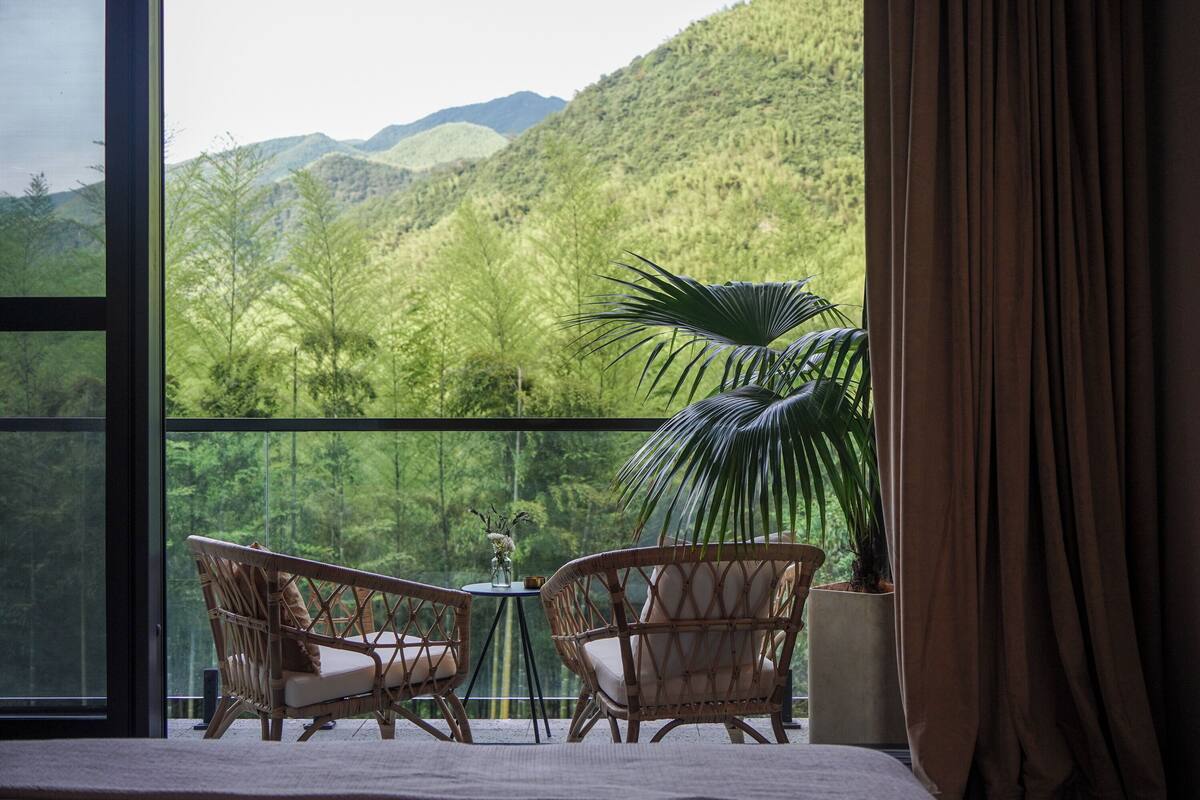 A balcony with wicker chairs and a table, overlooking a lush green landscape with mountains.