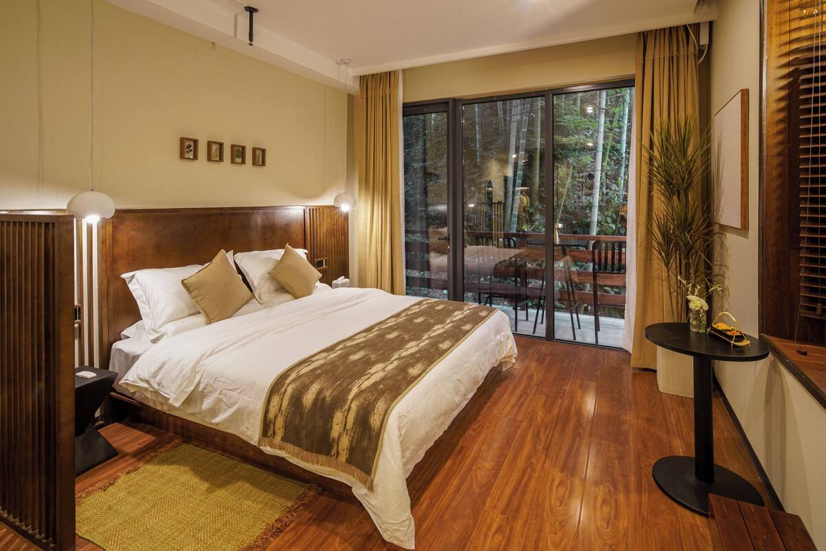A spacious bedroom with a large bed, wooden flooring, a balcony with a view of trees, and a small table with a vase of flowers.