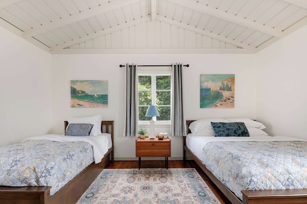 A bedroom with two beds, a window with curtains, and paintings on the walls.
