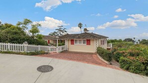 A single-story house with a red door and white picket fence, surrounded by greenery and a clear sky.