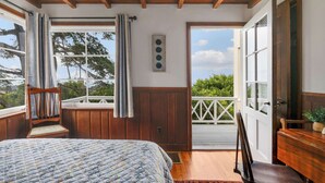 A bedroom with a wooden ceiling, a bed with a quilt, a chair, a window with curtains, and a view of greenery outside.