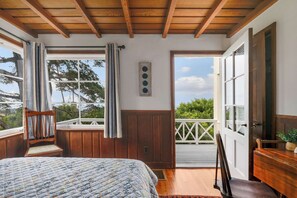 Un dormitorio con techo de madera, una cama con colcha, una silla, un ventanal con cortinas y vistas al exterior con vegetación.