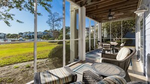 A covered patio with wicker furniture, large windows, and a view of a green field and houses.