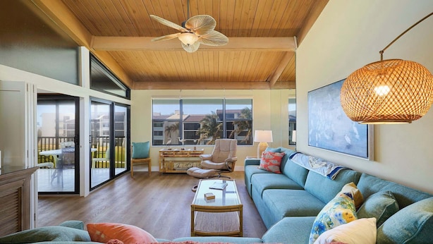 A spacious living room with a ceiling fan, a large window, a sofa, a coffee table, and a hanging light fixture. - Loggerhead 523 | Beachfront Condo with Vaulted Ceilings | Steps to the Sand (Sanibel)