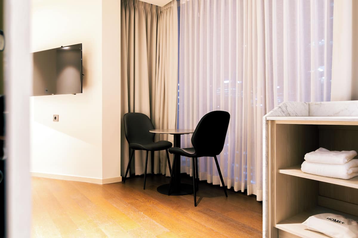 A hotel room with a flat-screen TV mounted on the wall, two black chairs around a small round table, a wooden floor, and a shelf with folded towels.