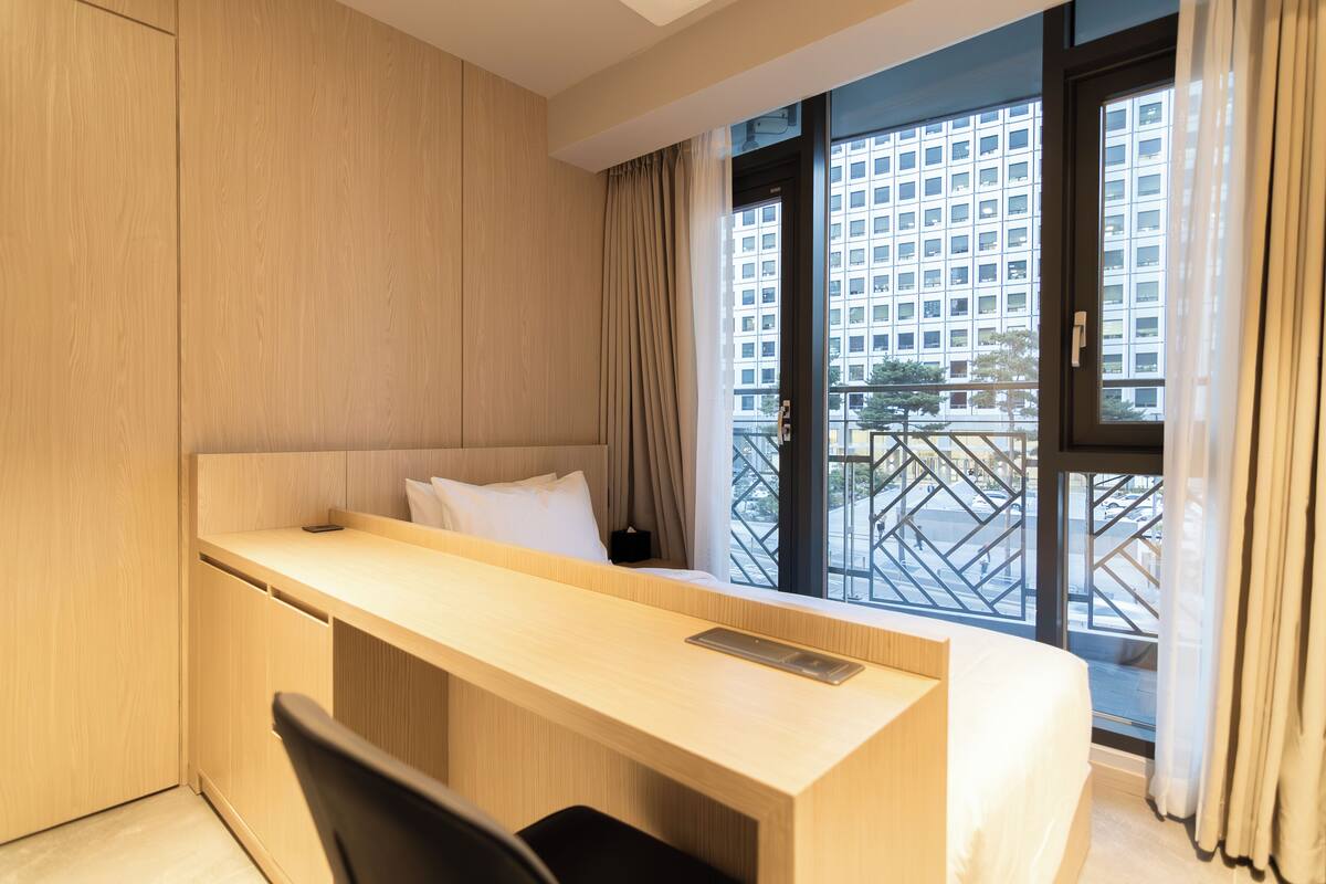 A hotel room with a wooden desk, a bed, and a large window with a view of a building.