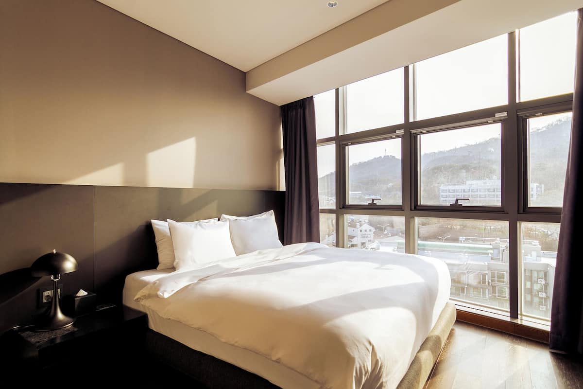 A hotel room with a large bed, a nightstand, a lamp, and a view of a cityscape through large windows.