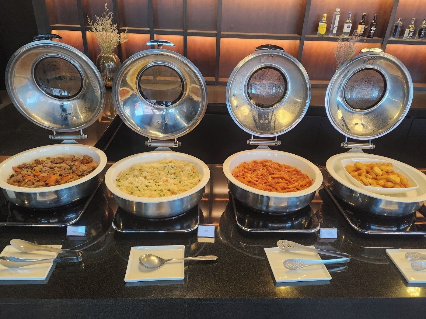 Four covered food dispensers on a counter with utensils and a small sign.