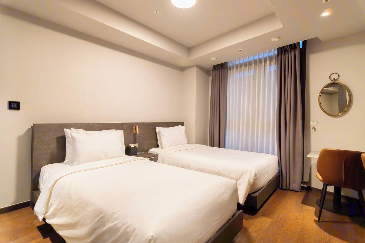 A hotel room with two beds, a desk, a chair, and a large window with curtains.
