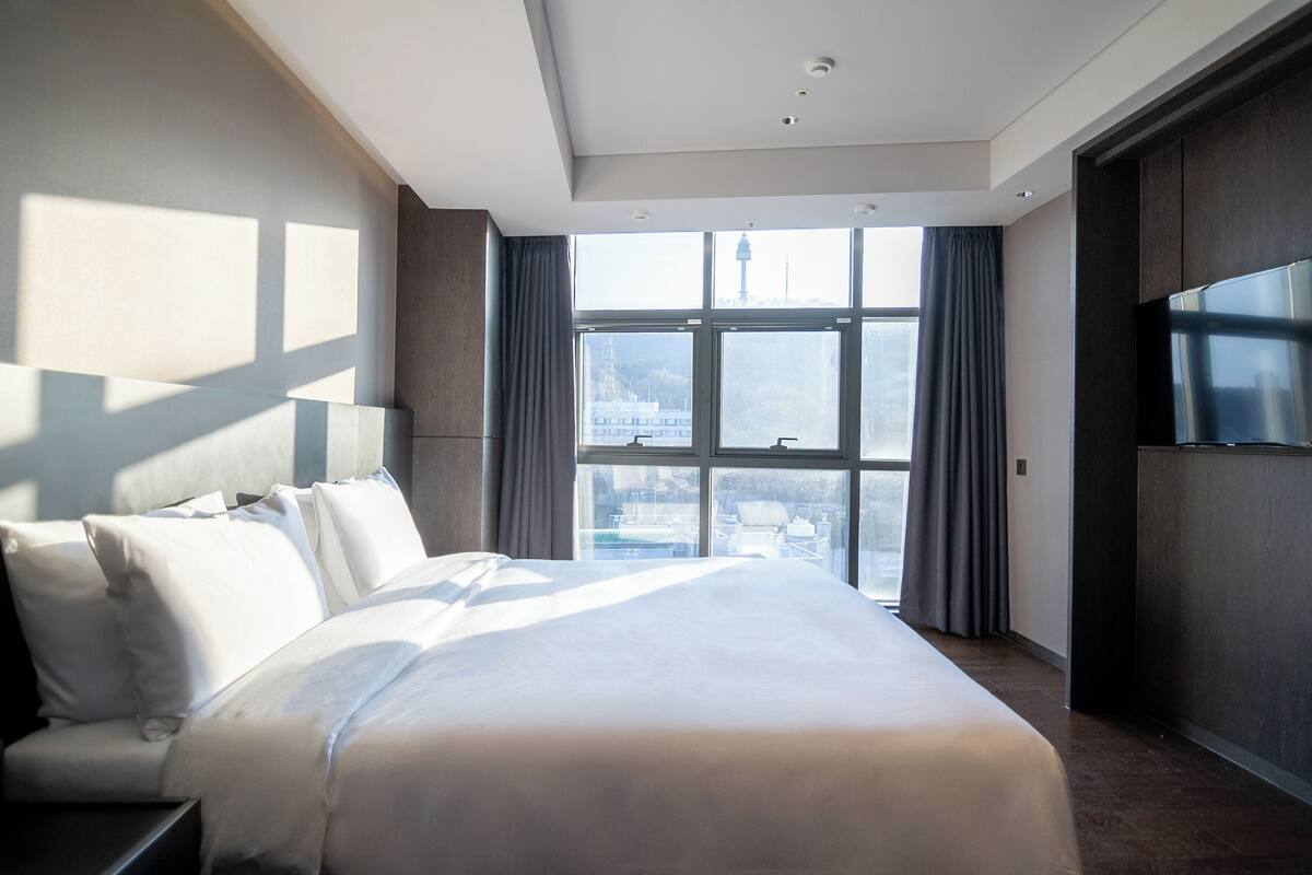 A modern hotel room with a large bed, a TV, and a city view through the windows.