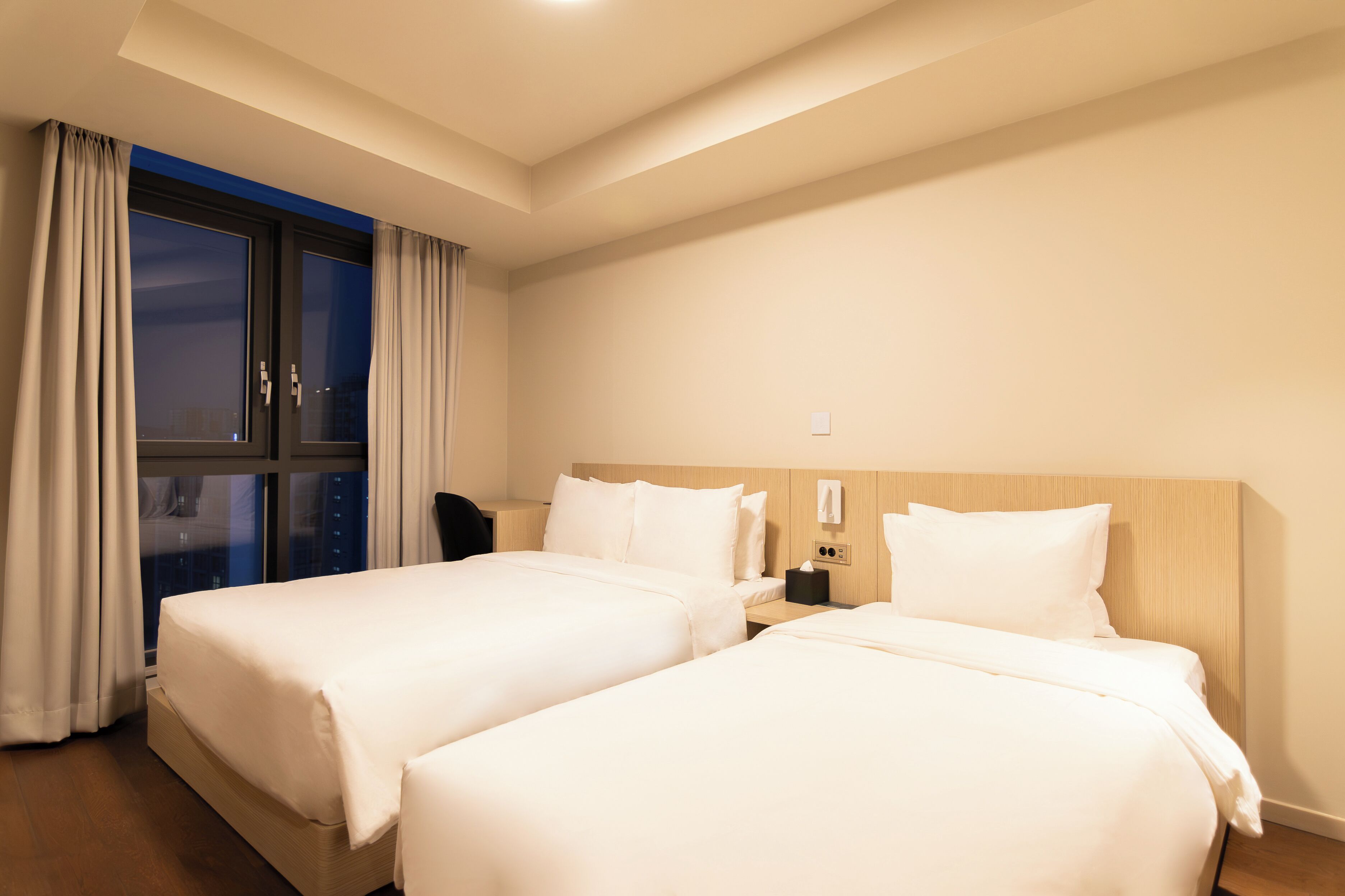 Habitación de hotel con dos camas, una mesita de noche, un escritorio y una ventana con cortinas.