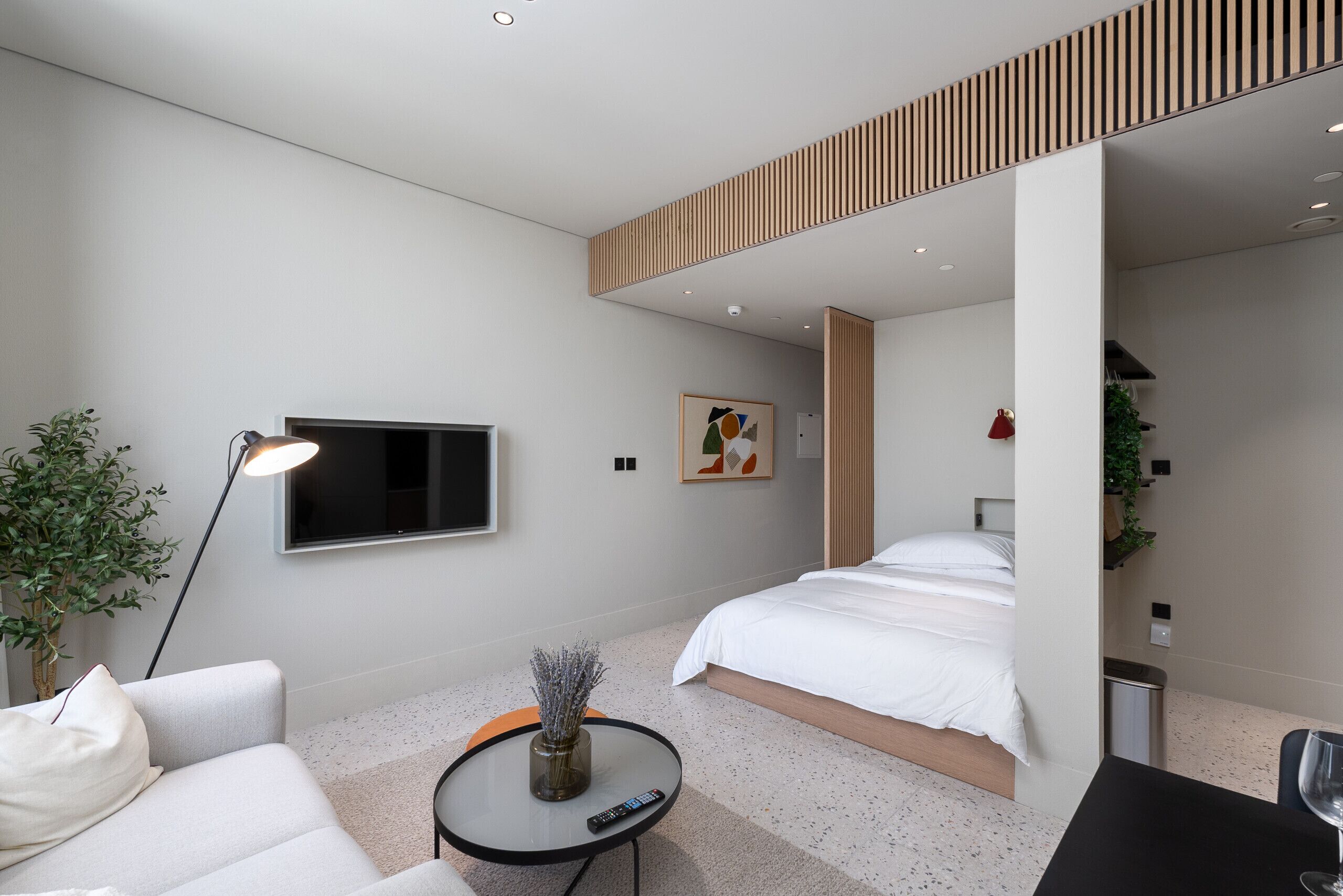 Ein modernes Hotelzimmer mit Bett, Sofa, kleinem runden Tisch mit Vase, Fernseher und Pflanze.