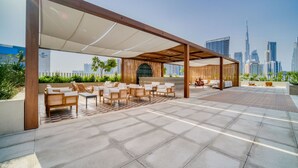 An outdoor seating area with multiple chairs and tables, a wooden pergola, and a city skyline view in the background. - Trendy Studio w/ Burj Khalifa View (Dubai)