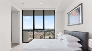 A bedroom with a large window, a bed with white linens, a dark headboard, and a framed picture on the wall. - Luxe 2-Bed: Modern Interiors, Balcony & BBQ Area (Lidcombe)
