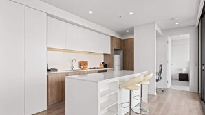 A modern kitchen with white cabinetry, a central island with bar stools, and a wooden accent wall. - Luxe 2-Bed: Modern Interiors, Balcony & BBQ Area (Lidcombe)