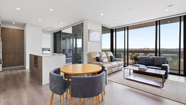 A modern apartment with a kitchen, dining area, and living room with large windows offering a city view. - Luxe 2-Bed: Modern Interiors, Balcony & BBQ Area (Lidcombe)