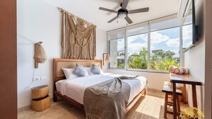 A bedroom with a bed, a ceiling fan, a large window, a wooden nightstand, and a small table. - NHOA 305B Lux 1BR Pool View Aldea Zama (Tulum)