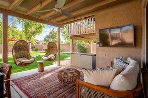 A covered patio area with wicker chairs, a sofa, a TV mounted on the wall, and a patterned rug.