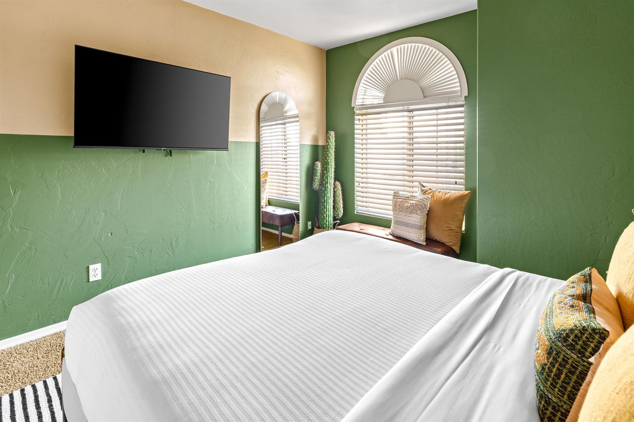 A bedroom with a large bed, a flat-screen TV mounted on the wall, and a window with blinds.