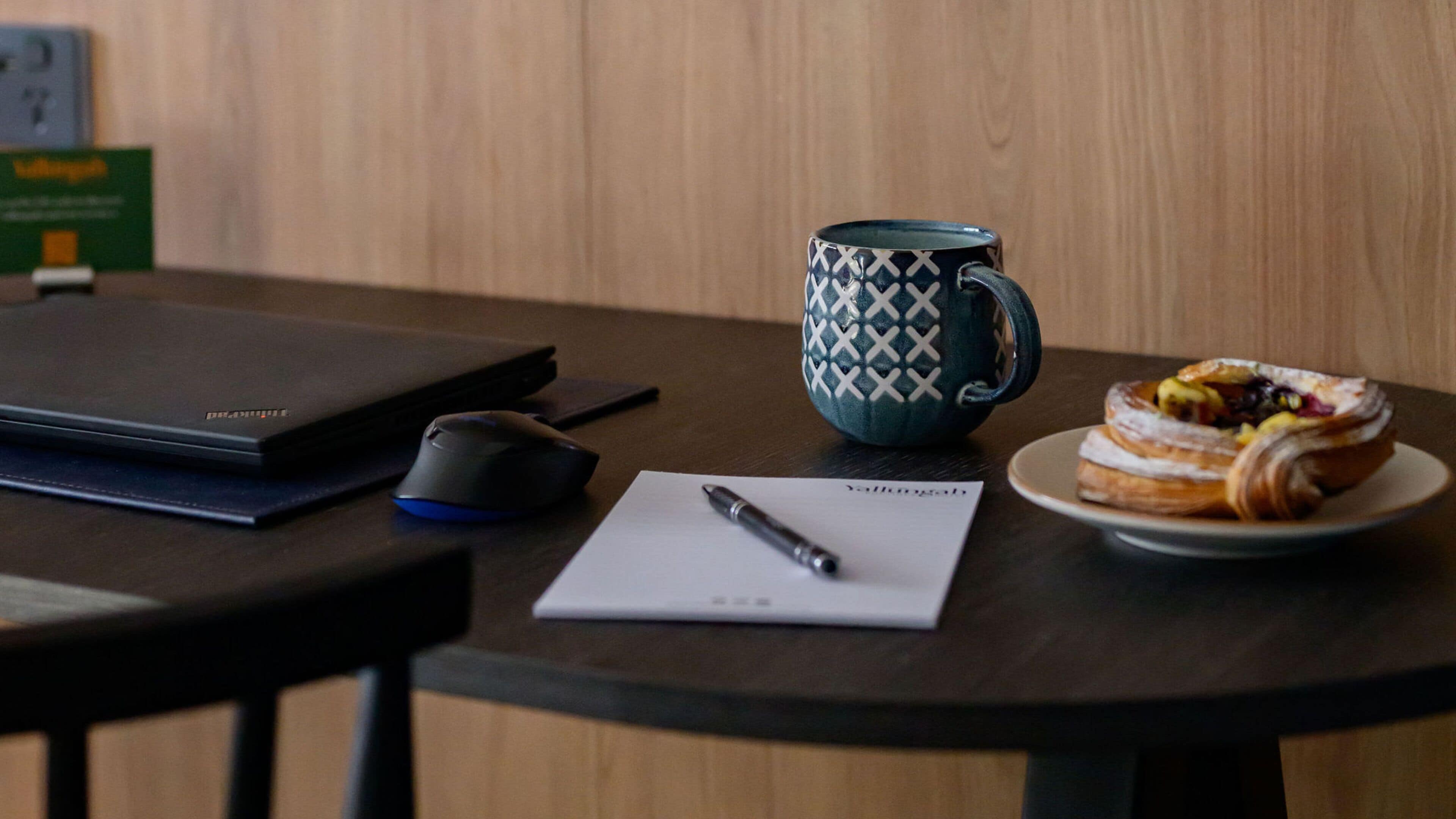 A laptop, a mouse, a mug, a pen, a notepad, and a plate with a pastry on a wooden table.