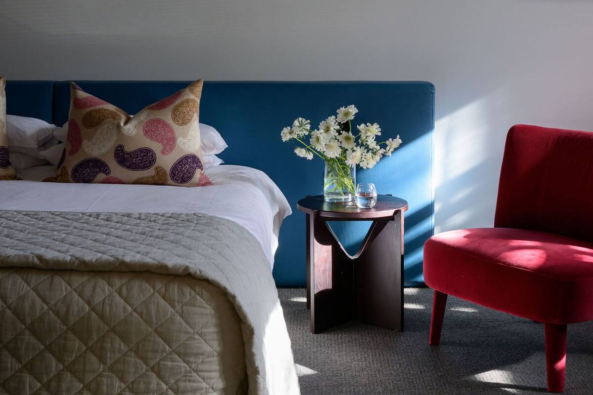 Una habitación de hotel con una cama, una mesita de noche con un florero y un sillón rojo.