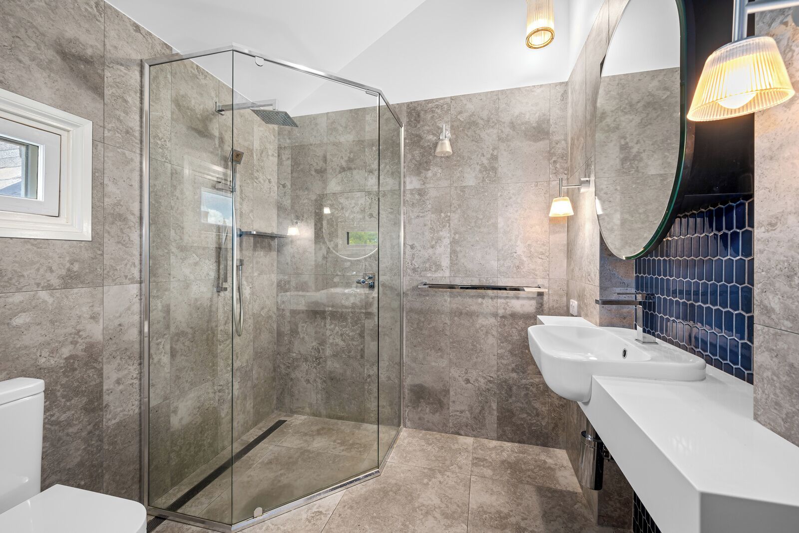 Een moderne badkamer met een glazen douchecabine, een wit wastafelmeubel en een betegelde spatwand.