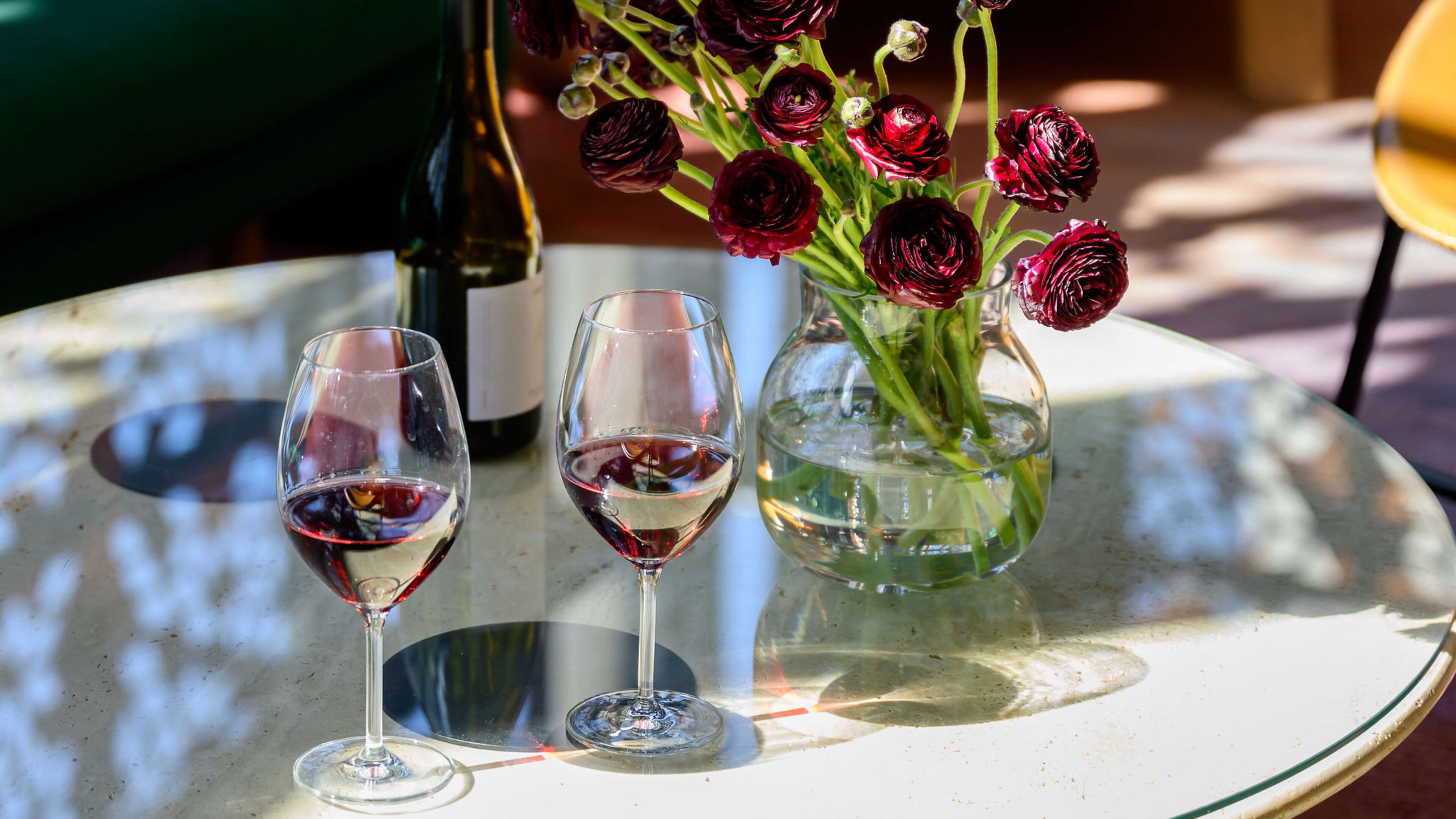 Two wine glasses on a round table with a bottle and a vase of flowers.