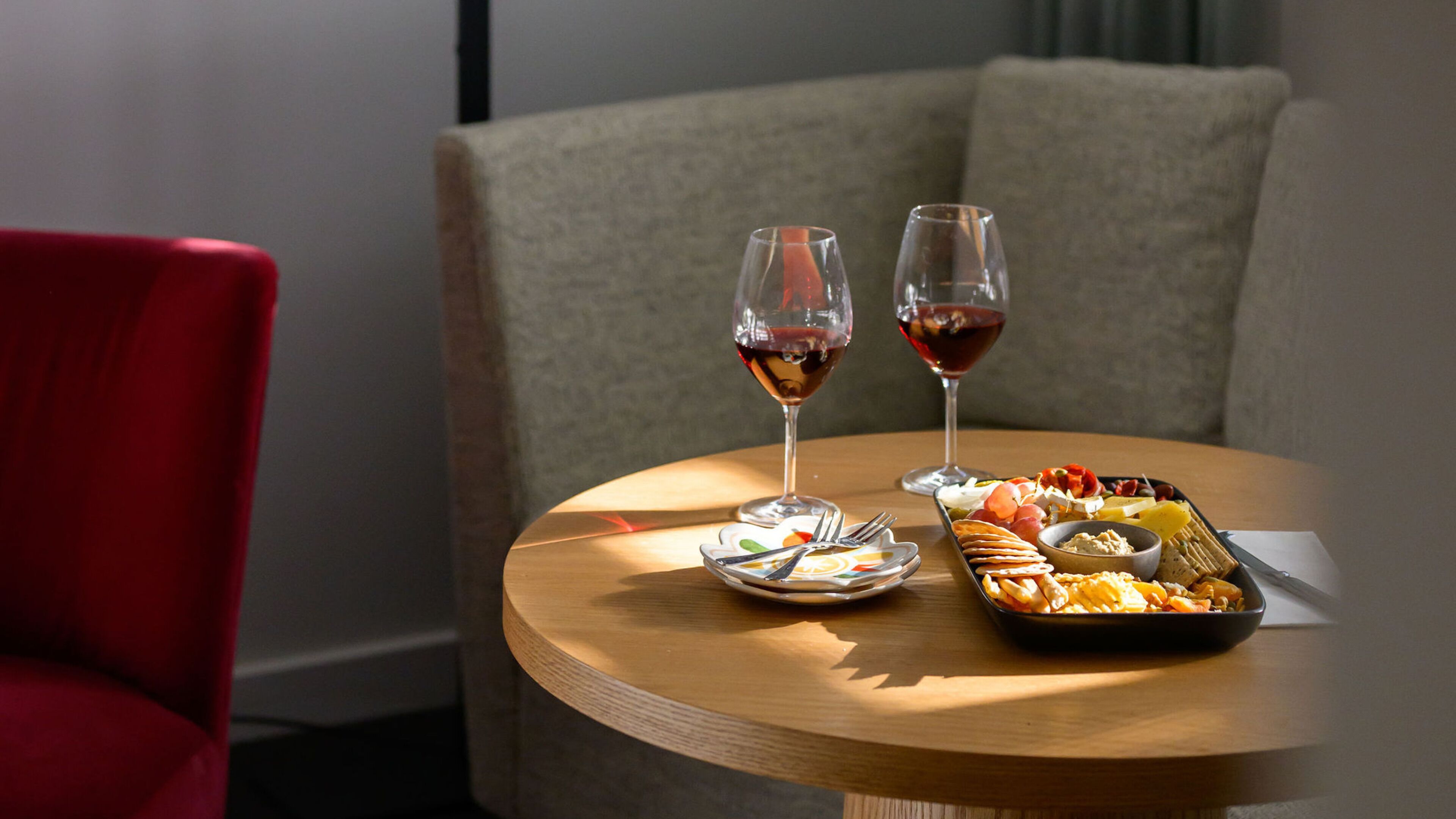 A round wooden table with a plate of snacks, two glasses of red wine, and cutlery.