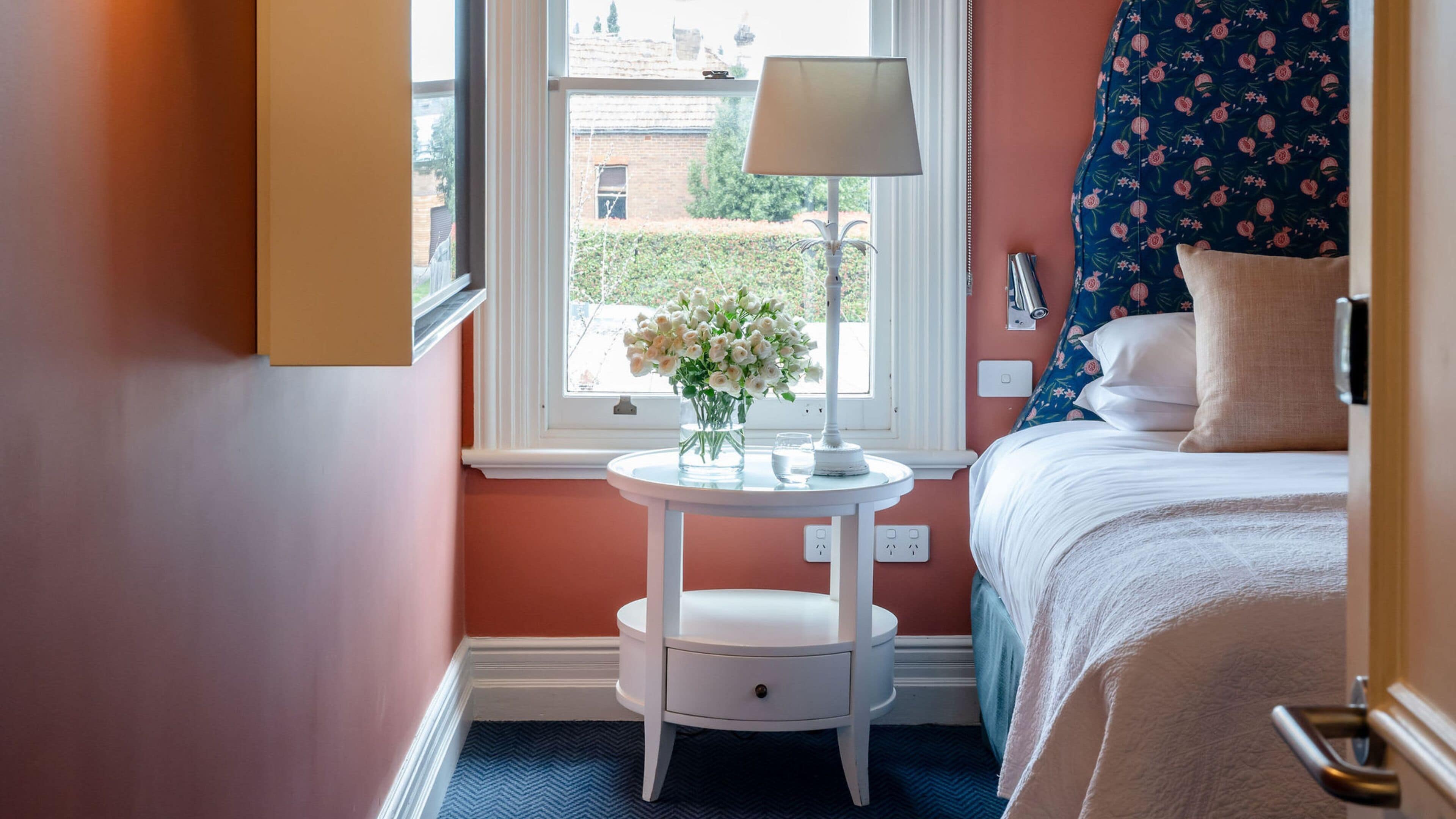 A bedroom with a bed, a bedside table with a lamp, a vase with flowers, and a window with a view of the outside.
