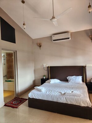 Une chambre d’hôtel avec un grand lit, un ventilateur de plafond, un appareil de climatisation, une petite table de chevet avec une lampe, et une vue sur l’espace de la douche.