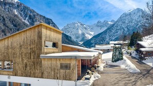 Una cabaña de madera con techo nevado, fachada de madera y vistas a montañas nevadas.