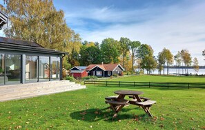 A modern house with large glass windows, a wooden deck, and a picnic table in a grassy area with a lake view.