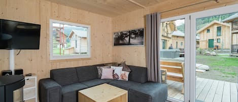 Un salon moderne avec un intérieur en bois, un canapé gris, une table basse en bois, un téléviseur à écran plat et une grande porte-fenêtre coulissante donnant sur une terrasse extérieure.