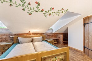 A bedroom with a wooden bed, a nightstand, a wall-mounted lamp, and a sloped ceiling with floral decoration. - Room 'Fuchswirt 01 - Doppelzimmer' with Mountain View, Shared Garden and Wi-Fi (Kelchsau)