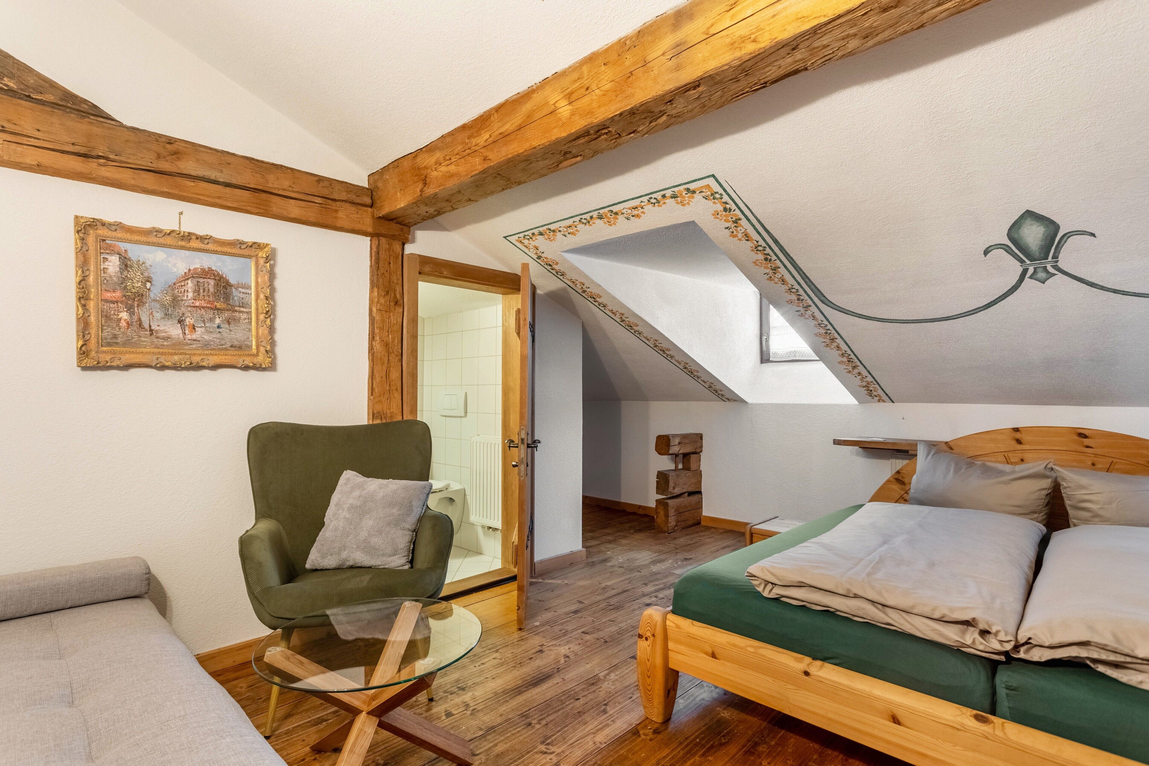 Een kamer met een houten plafond, een bed met groene beddengoed, een groene fauteuil, een glazen tafel en een ingelijst kunstwerk aan de muur.