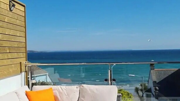 Un balcón con un sofá blanco y cojines naranja, con vistas a un mar azul cristalino y una costa lejana.