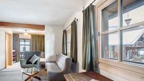 Ein Zimmer mit einer Couch, einem Sessel, einem Holztisch und großen Fenstern mit Vorhängen.