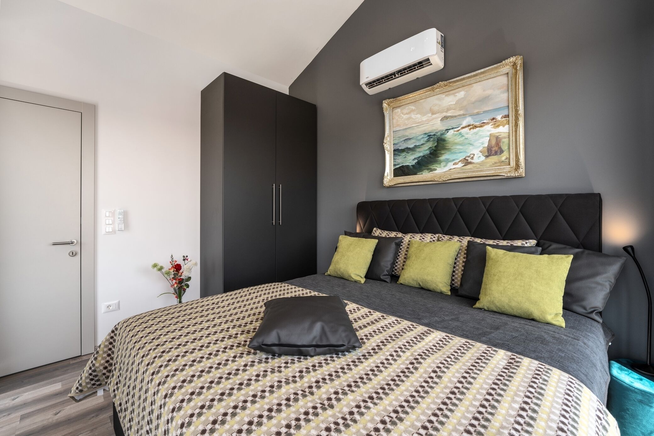 A modern bedroom with a large bed, a dark headboard, a painting, and an air conditioning unit.