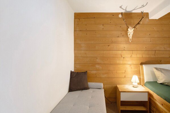 A small, cozy room with a wooden ceiling, a bed with a green mattress, a nightstand with a lamp, and a mounted deer skull.