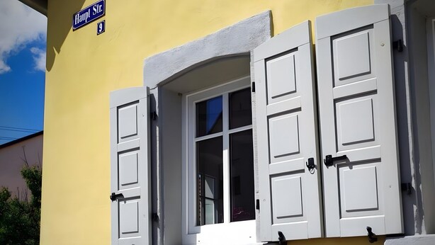 A building with a yellow facade, a window with white shutters, a stone base, and a street sign reading 'Haupl Str. 9'. - Apartment 'La Maison Palatinate' with Charm, Southern Wine Route (Kapsweyer)